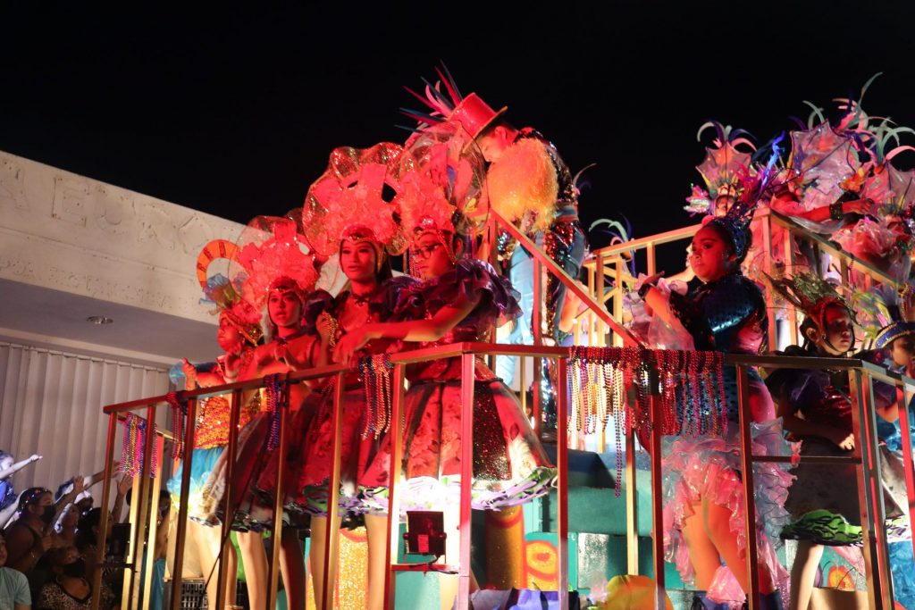 A group of costumed performers along a railing of a float they are riding in a Cozumel Carnaval parade. They appear to be wearing red dresses with full skirts and large feathered headpieces.