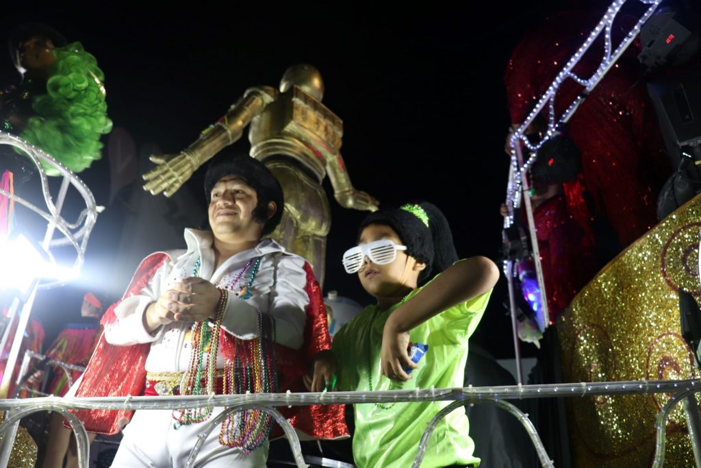 An Elvis costumed man throws mardi gras bead necklaces to the crowd from a float. There is a child standing next to him and a Star Wars costumed golden robot character is in the background.