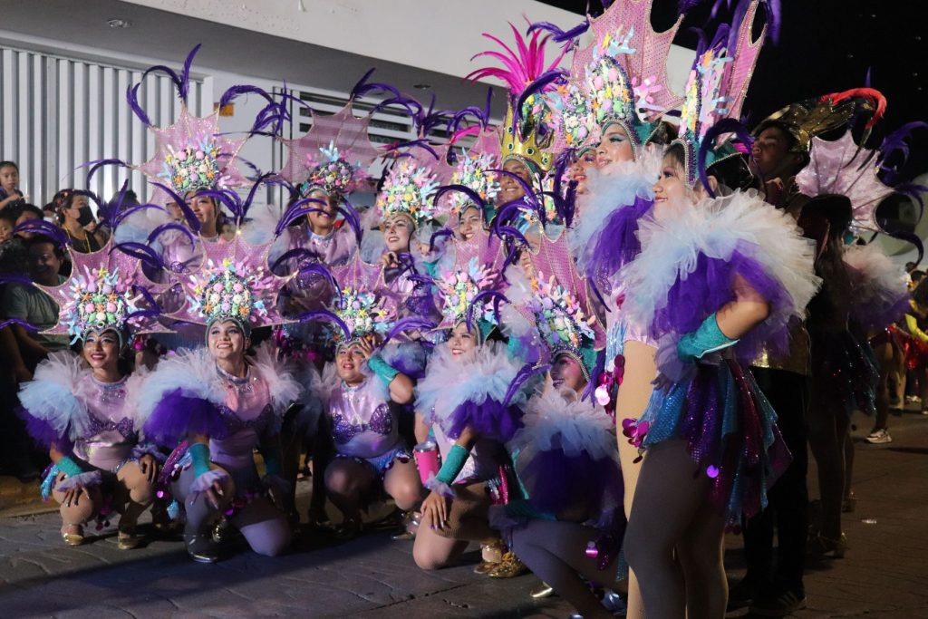 Female dance troupe poses along the Carnaval parade route in Cozumel. They are wearing white and purple costumes with large silver and purple headpieces.