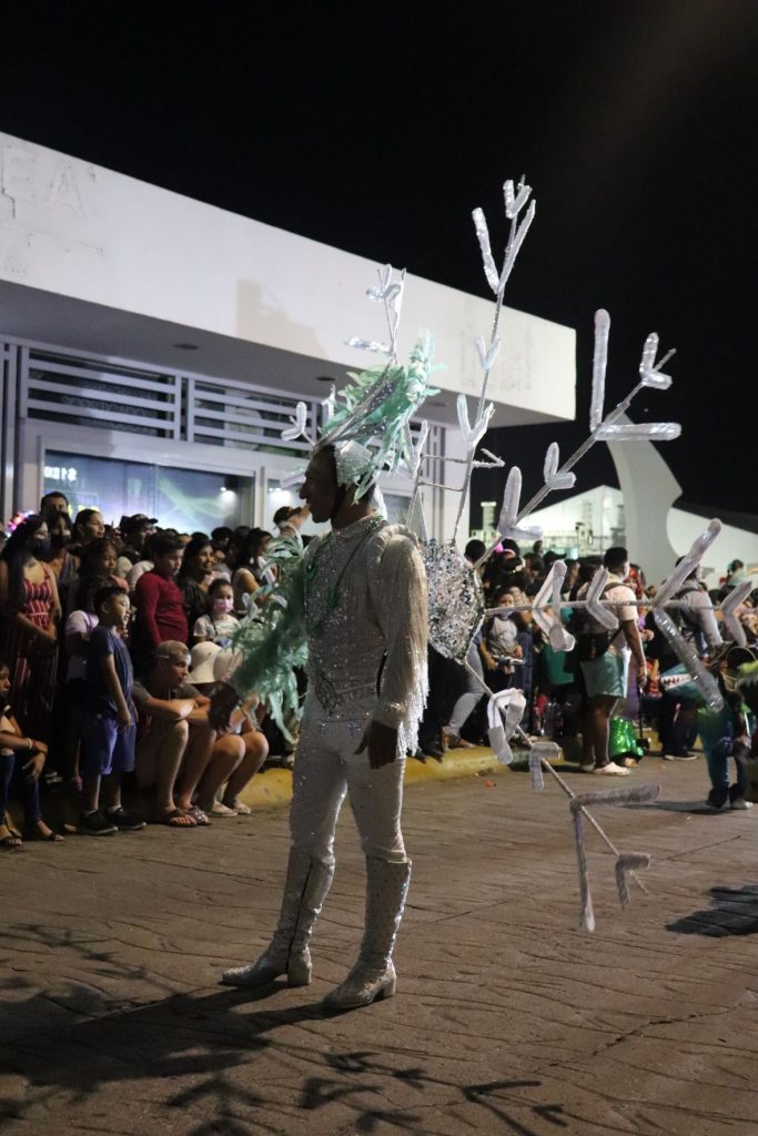 Spectators line the street as a Cozumel Carnaval performer walks by. He is wearing a large white irredescent snowflake costume with a 7 foot wide back piece and a tall feathered headpiece. He is dressed in a white and silver long sleeved costume and wearing knee high boots as he walks the parade route.