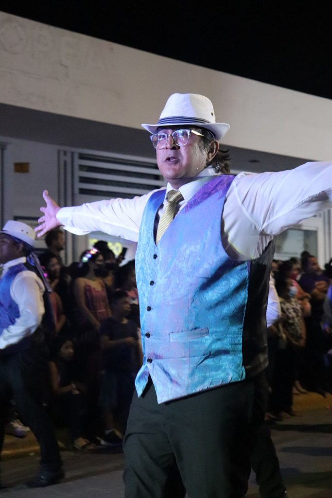 A middle aged latino man wearing a fedora and purple vest with his arms wide spread dances his way along the Cozumel Carnaval parade route.