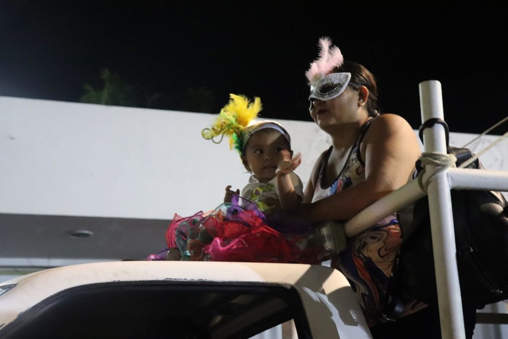 Mother and toddler daughter, both dressed in festive masks and headdresses for Carnaval in Cozumel ride on top of a small float during a parade.