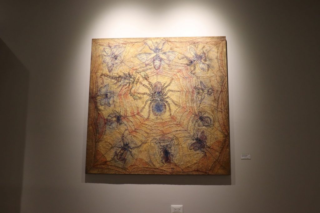 Large painting on a wall mostly done in brown tones. There is a large spider in the center with a web extending across the entire canvas. There are a variety of flying insects that appear trapped in the web.