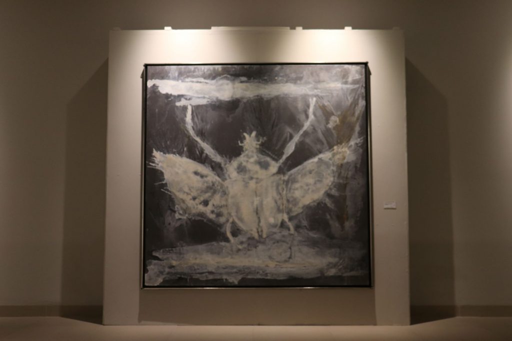 A large painting displayed on a wall. The art is done in gray and cream and appears roughly like a winged insect.