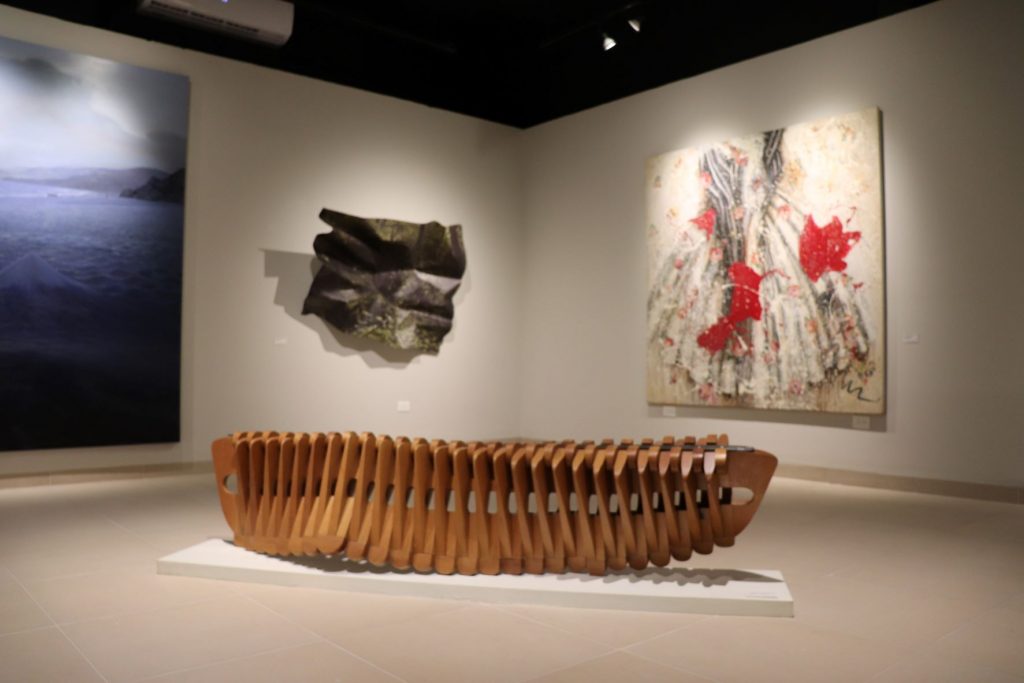 Large and low wooden sculpture in the middle of a gallery room with several pieces of art displayed on the walls.