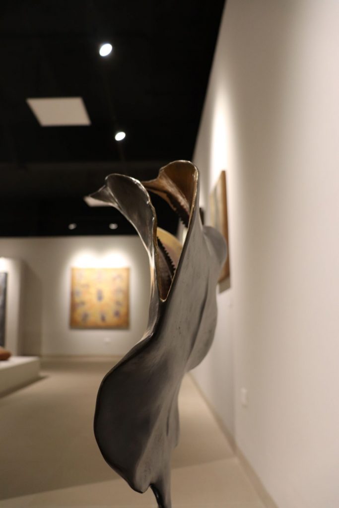 Closeup bronze modern sculpture with a large painting shown mounted on a wall in the distance.