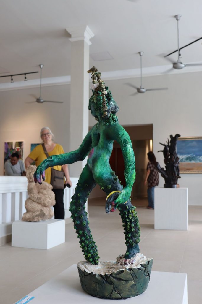 Modern green sculpture of a sea creature/merman in gallery in Cozumel. He stands on a base of sand and leaves and holds a tropical fish in a hand.