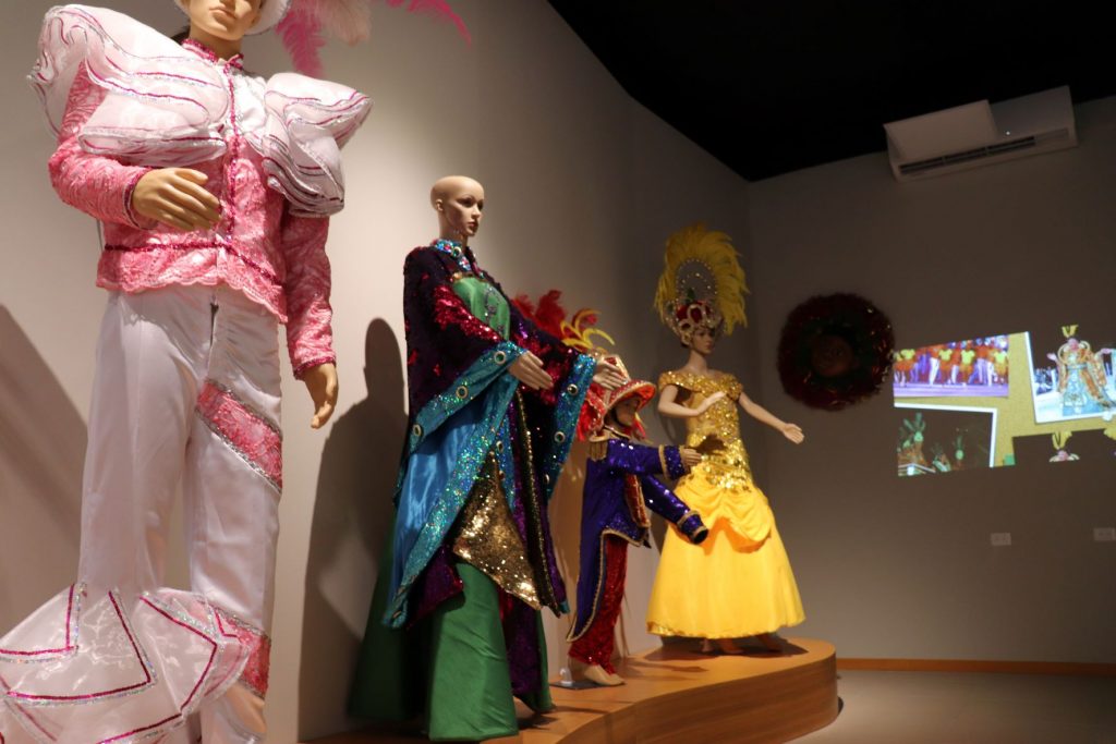 Manequins wearing actual Carnaval costumes on display in the second floor Cozumel Carnaval gallery. There is a video projected on a side wall that shows the history of Carnaval on the island. A men's elaborate white and pink outfit, a child's purple and red outfit and a woman's long yellow gown are shown.