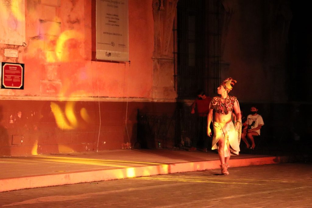 A Latino man dressed in Mayan attire is cast in a projected golden light as he looks at the Cathedral that towers over him.