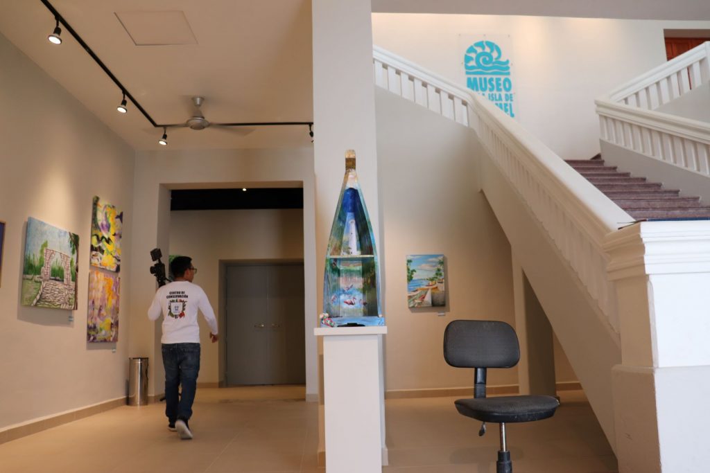 View from the front door and lobby of Museo de le Isla de Cozumel shows the grand staircase to the second floor and pieces of art on display on the first floor. There is a man visible from the back wearing a Cozumel staff long sleeve t-shirt. The museum logo is in aqua blue mounted to the wall above the stairs, so it is visible when you first enter the building.