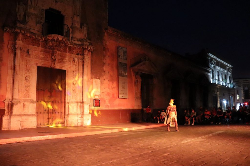 A landscape view shows a Latino man dressed in authentic 16th century Mayan attire. He walks in front of the crowd, eyeing a projected pattern that moves about on the Cathedral walls.