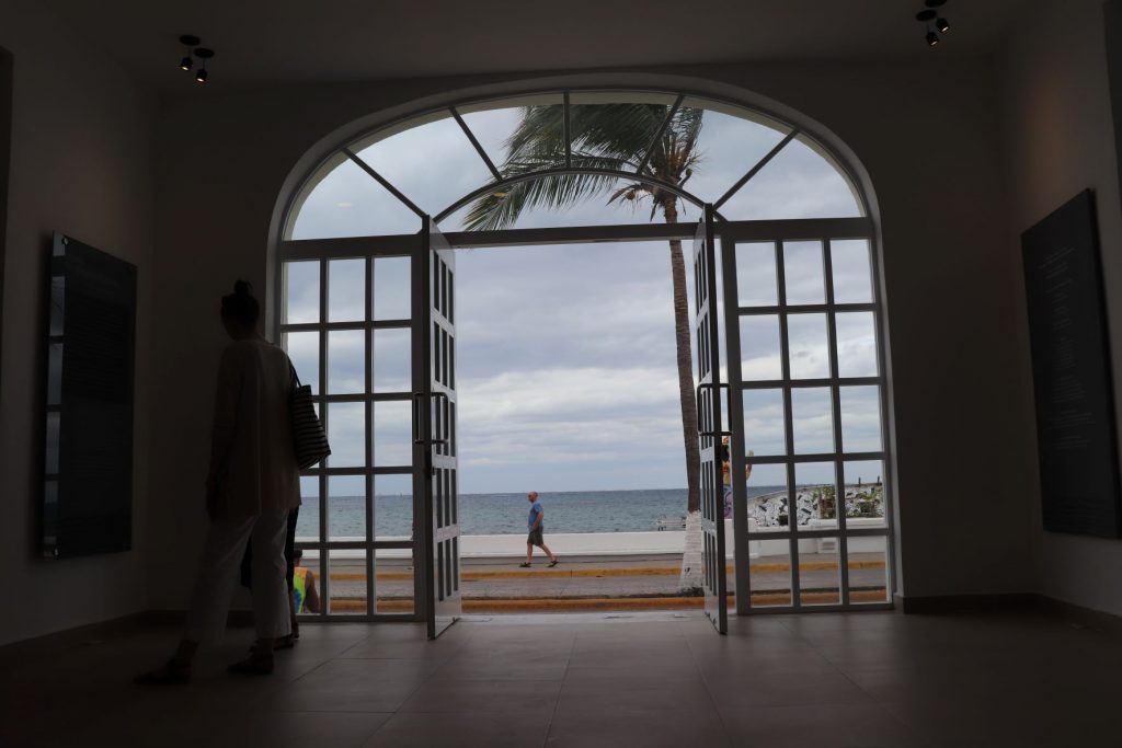 The view of the ocean across the street from the museum, when looking out the open front doors. There is a man walking along the malecon and a tall palm blowing in the breeze. In the shadows inside the museum,, you can see a woman studying the art on the inside. The main entrance is an arch the width of 4 large doors. The side windows, doors and upper arch are all divided into smaller window panes with crisp white paint.