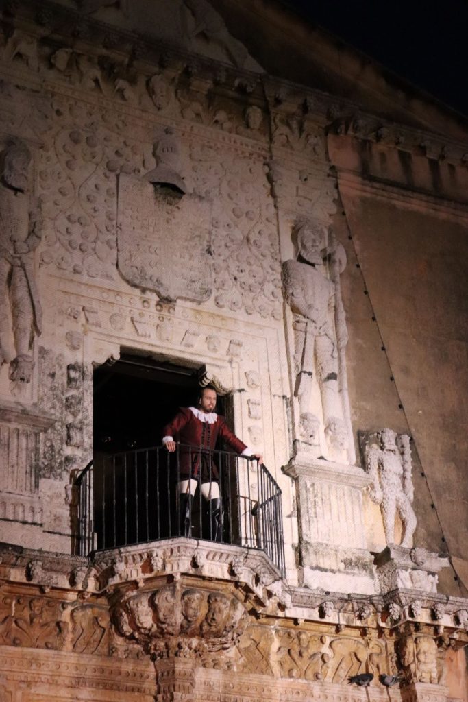 A man dressed as a 16th century conquistador stands on the Cathedral balcony, his hands gripping the back bar in front of him. He stares down at the crowd sternly.