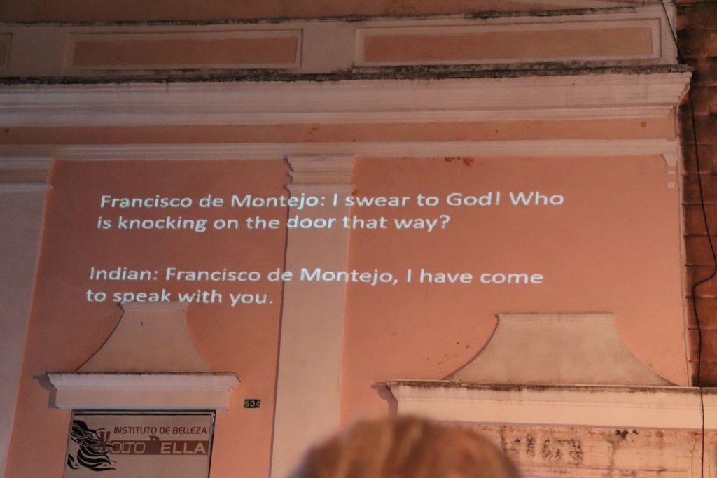 The projected on the pink Cathedral building are white and large. They are in English to accommodate those who do not speak Spanish. Francisco de Montejo is the first speaker, saying: "I swear to God! Who is knocking on the door that way?" The next speaker, entitled "Indian", says: "Francisco de Montejo, I have come to speak with you."
