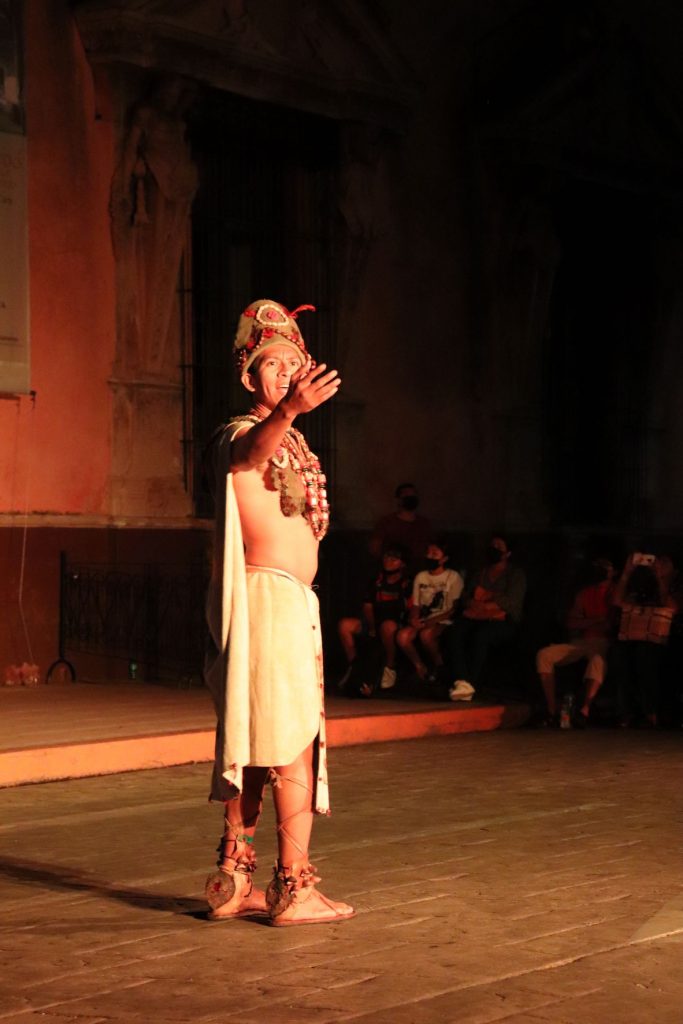 A portrait view of a Latino man is seen. He is dressed in authentic, 16th century Mayan garb as he stands, his arm out, speaking to the crowd. His face is stern and serious as he focuses on his message.