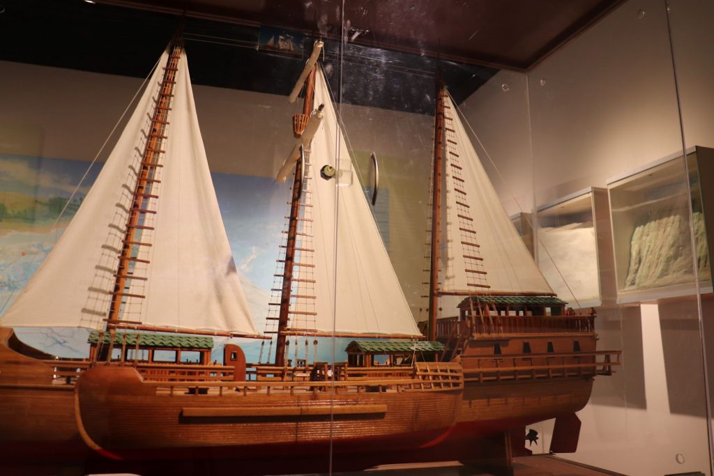Three masted wooden sailboat model on display. This would have been a huge sailboat similar to the way you might think of a old pirate ship. Medium dark wood. Cream fabric sails and sections of boat covered in green tiles roof. This piece is delicate and kept enclosed in glass cabinet at Museo de la Isla de Cozumel.