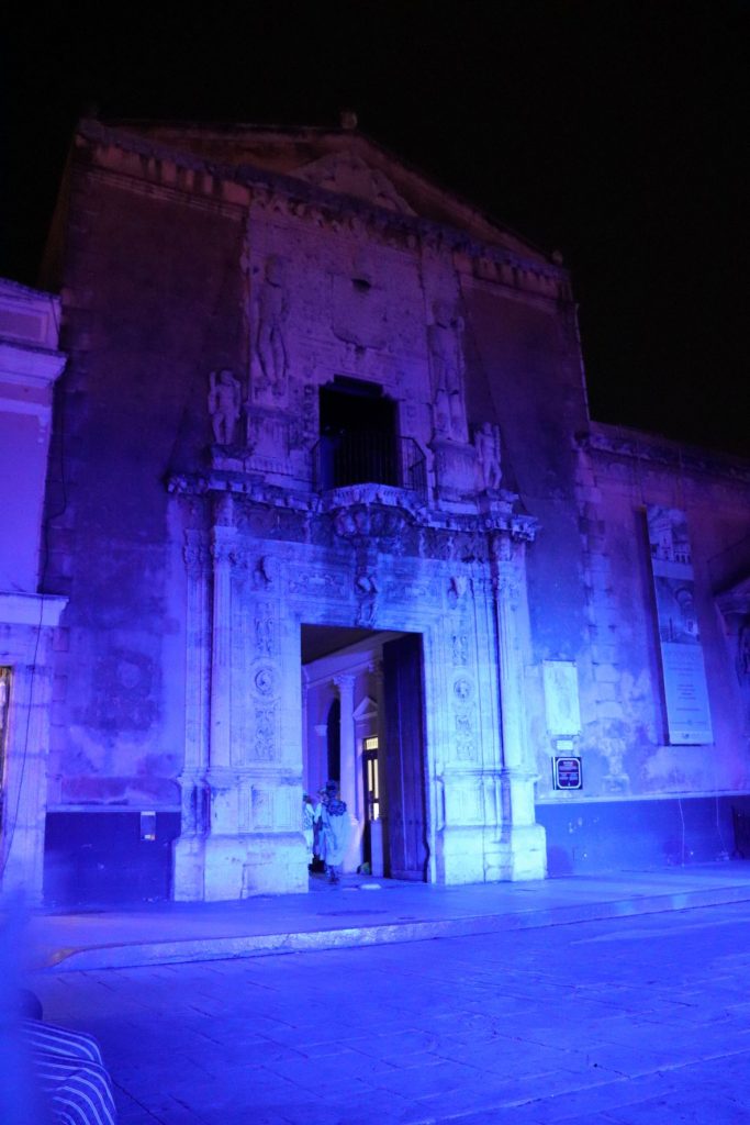 A view of the Cathedral is seen. Cast in ghoulish blue light, the building's two conquistador statues near the top make for an ominous sight. An empty, dark balcony juts out above the tall doorway. In the doorway, people can be seen beginning to walk out.