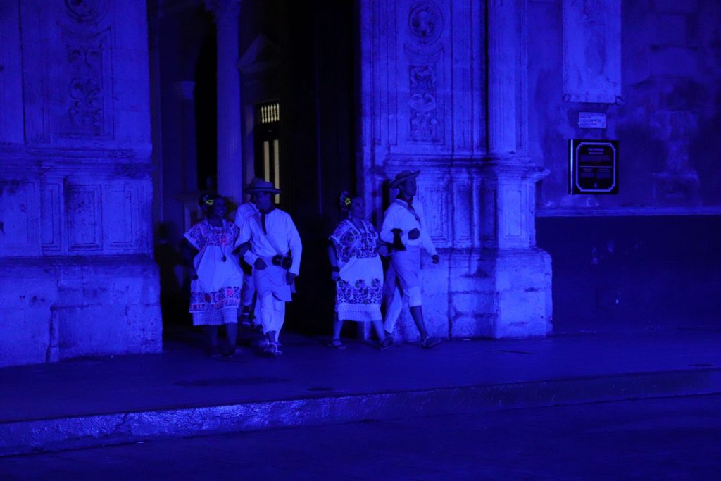 Two pairs of male and female Latino dancers slowly walk out, their arms interlocking. The men wear hats while the women hold flowers in their hair. The stage is set in a dark blue light. The inside of the Cathedral in which they exit can be seen only slightly; the elongated windows are visible while the Cathedral doors are open.