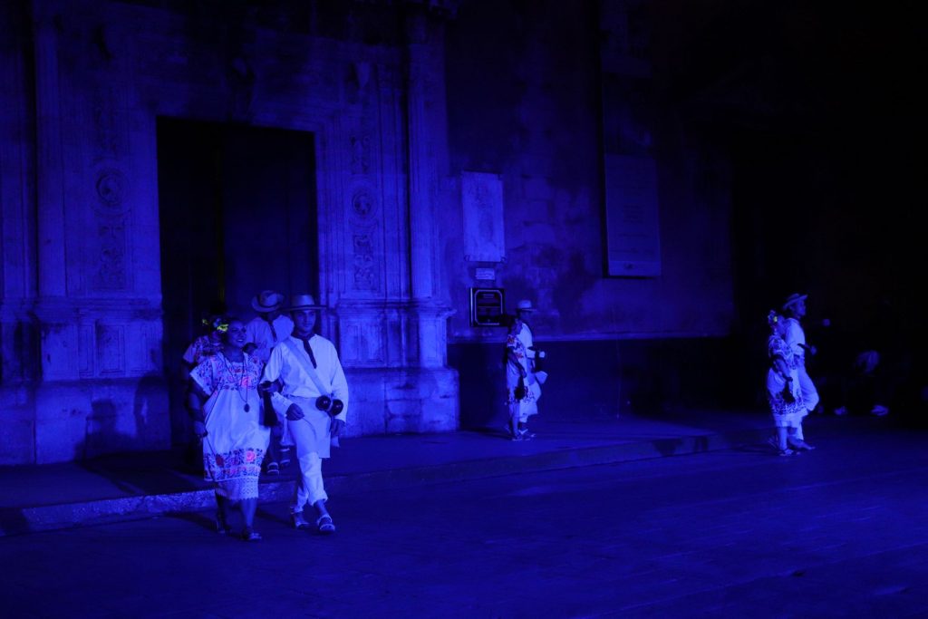 A landscape photo shows the stage which the Latino men and women dance upon. A dark blue light is cast upon the dancers and the stage. The men and women interlock their arms as they walk toward the hidden crowd. Behind them is part of the huge Cathedral in the town square. The building is adorned with a projection showing intricate patterns.