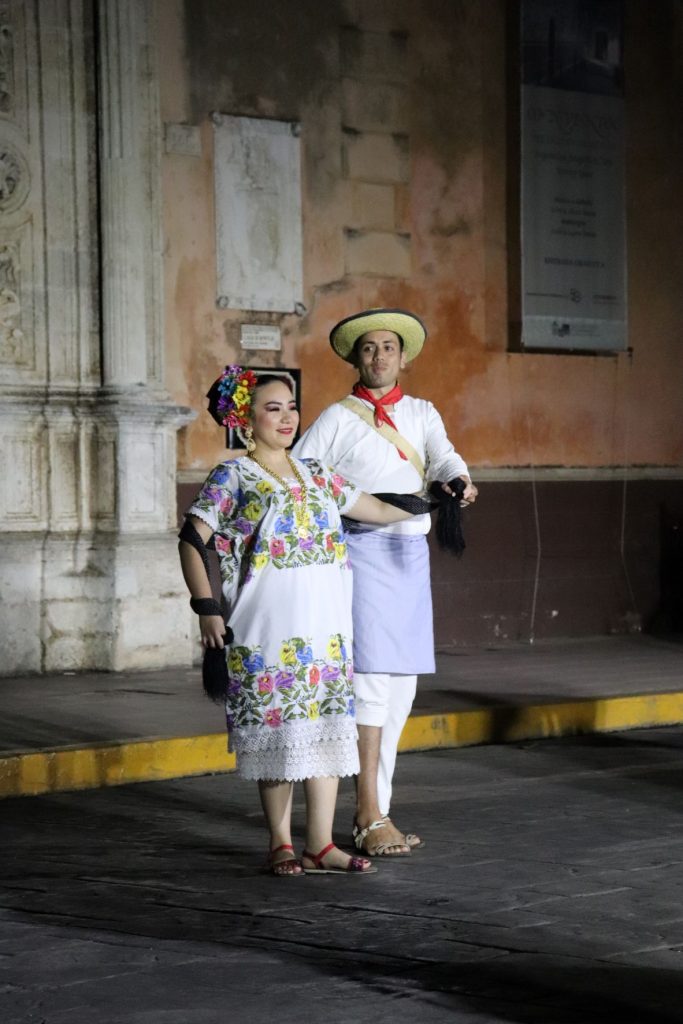 A portrait photo shows a Latino man and woman standing together. The man, wearing a hat and a red scarf, stands slightly behind the woman, who wears a floral dress and flowers in her hair. The woman wears a cheerful smile on her face while the man seems to be lost in pensive thought.
