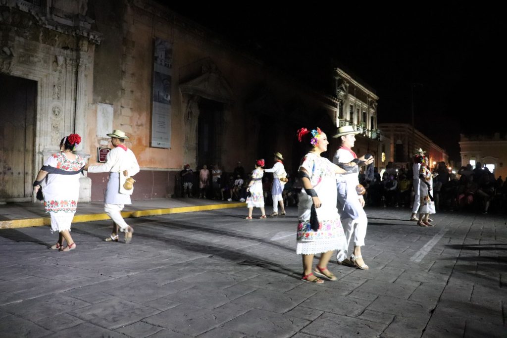 Four pairs of Latino men and women dance closely together. The men now wear their hats while the black sash wrap around the women's arms.