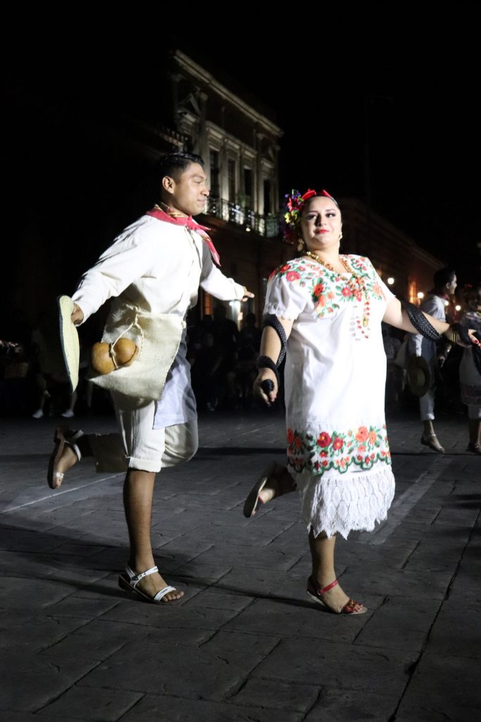 Two Latino dancers, a man and a woman, dance together. The man looks at the woman with a soft smile while the woman looks off onto her right with an equally small smile. The woman has a black sash laced around her arms while the man dons a sack tied around his waist.