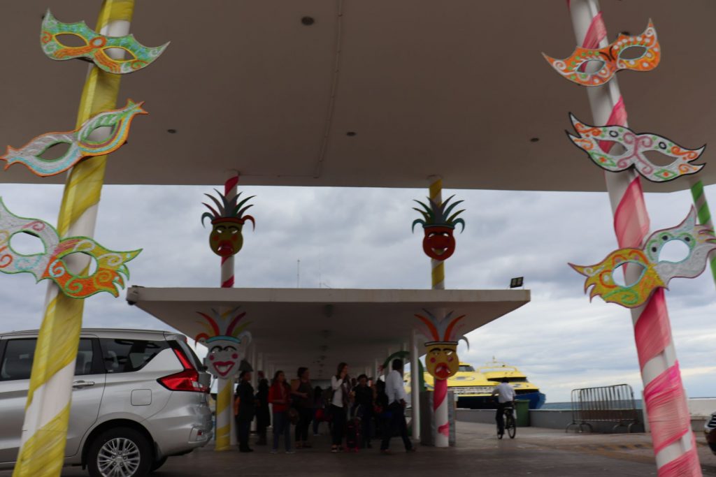 The entrance to the main ferry dock on Cozumel is decorated for Carnaval. Bright colors wrap the pillars like candy sticks and there are brightly colored Carnaval masks mounted on the pillars as well. To the right of the terminal,, you can see two bright yellow ferries docked.