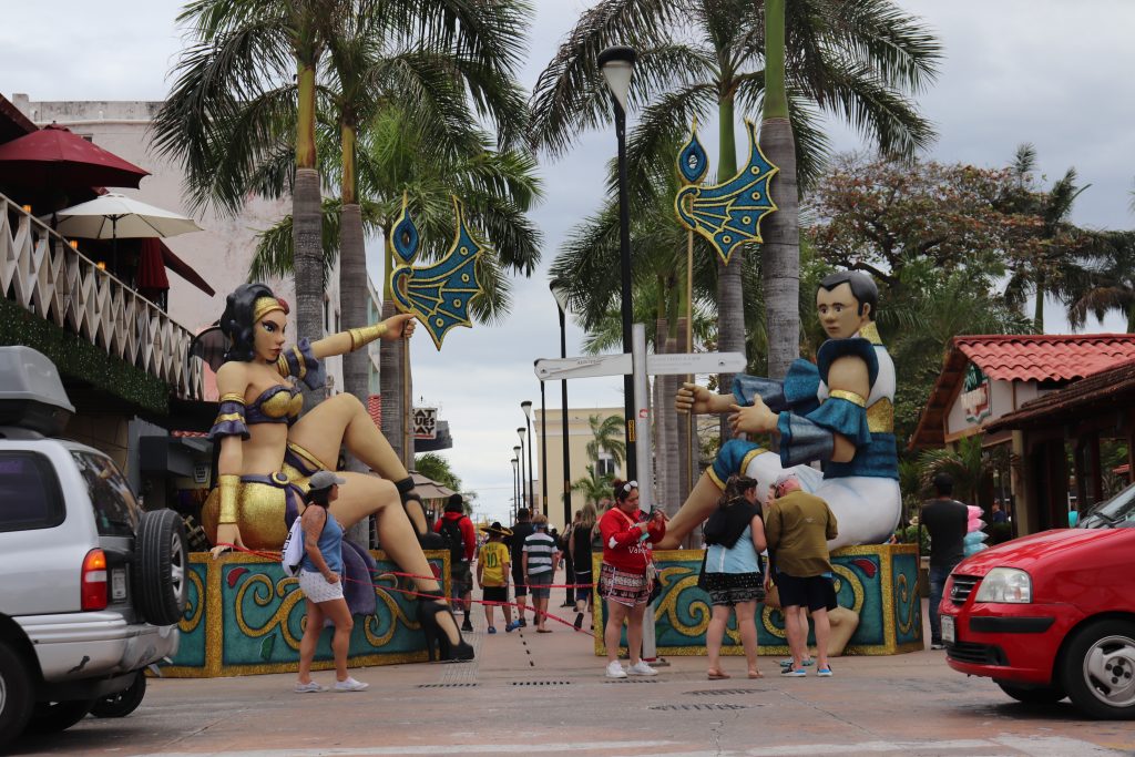 Very large display pieces for Carnaval season set at the entry to Benito Juarez park in Cozumel. These are more than twice the height of tourists standing near them. They are sculptures of costumed Carnaval performers, with a woman on one side and a man on the other.