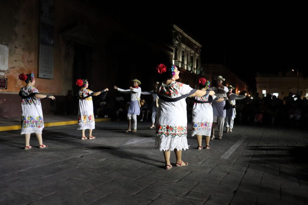 A group of Latino men and women dance together. Most face and look at each other, yet one female dancer looks at the crowd. The flowers in the women's hair are large and colorful.