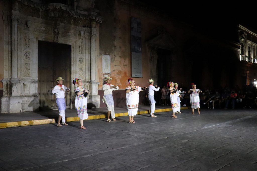 A line of eight Latino dancers, four male dancers and four female dancers all dance synchronously on the open-air stage. The men wear tan hats while the women wear white dresses with floral prints on them.