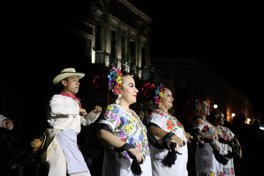 Four Latina female dancers stand beside each other while a Latino male dancer begins a dance move behind them. They all wear traditional Mexican clothing.