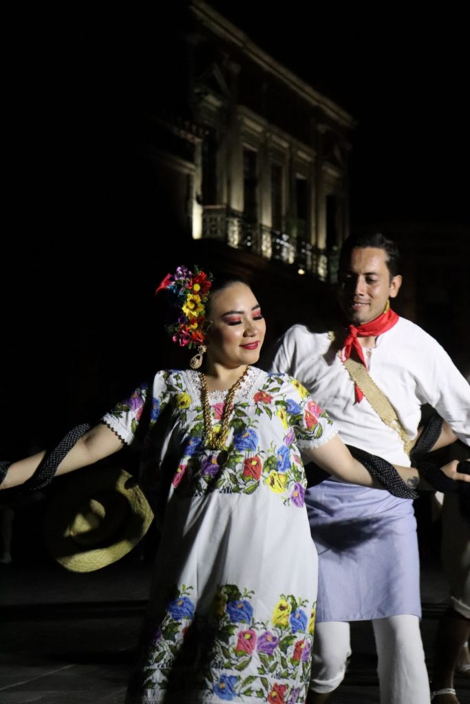 Two Latino dancers, a man and a woman dressed in authentic Mayan attire, dance closely together on the stage. The man wears a white shirt with a red scarf tied around his neck. The woman wears jewelry on her neck and an array of flowers in her hair.