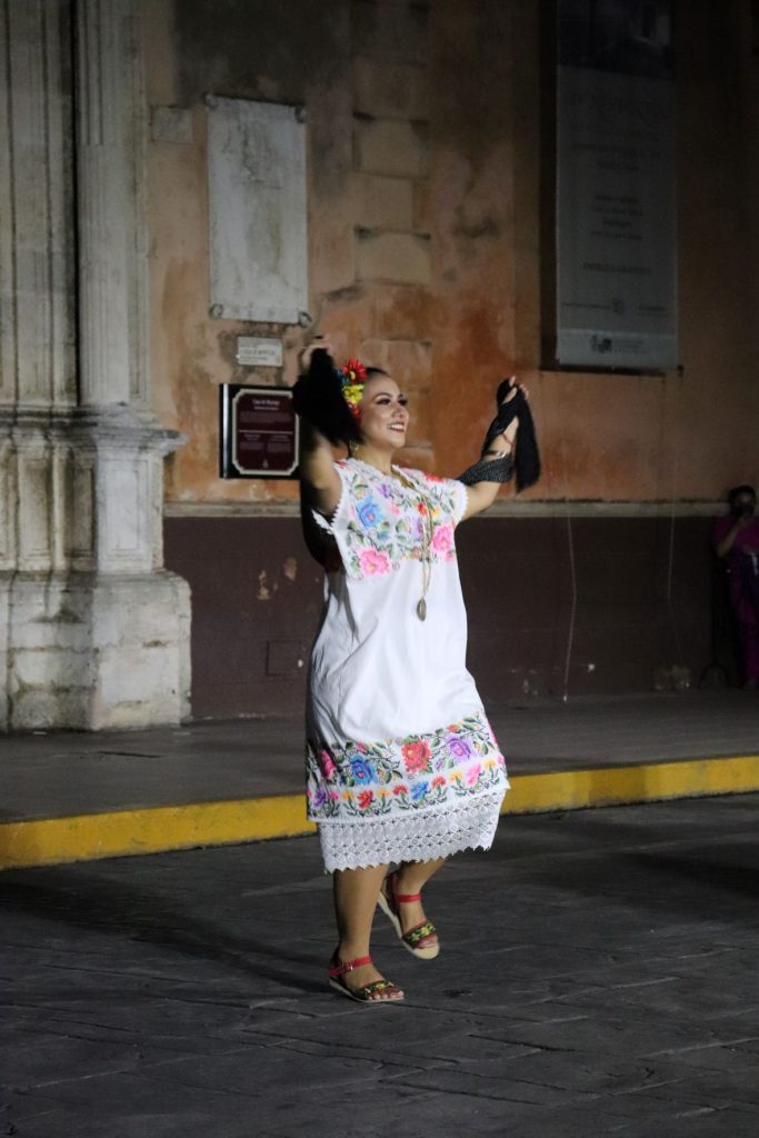 A Latina female dancer wears a traditional Mayan embroidered dress as she spreads her arms wide, dancing along to the music.