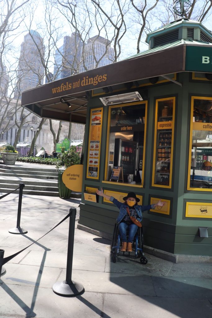 Anna, a teen girl sitting in her wheelchair is gesturing at the Wafels and Dinges kiosk behind her in the NYC park. She is wearing jeans and a denim jacket with a black fedora and yellow lower face mask. The kiosk is a muted green with yellow accents and signage.