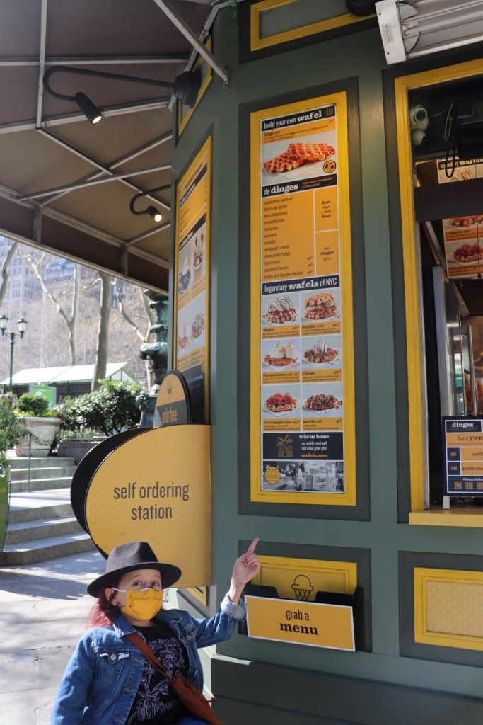 Anna, a teen girl using a wheelchair and wearing a black felt fedora,, points at the menu posted on the Wafels and Dinges kiosk in the park. You can see steps, bushes and a statue in the background.