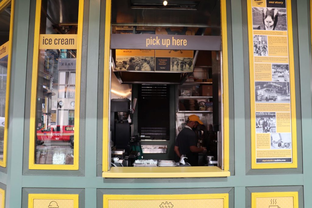 A view of the Wafels and Dinges kiosk with the open pick up window in the center, signs on the right and a banner for ice cream on the left. You can see into the NYC park kiosk through the window, and there is stainless steel kitchen equipment with a staffer wearing a yellow ballcap while working in the rear.