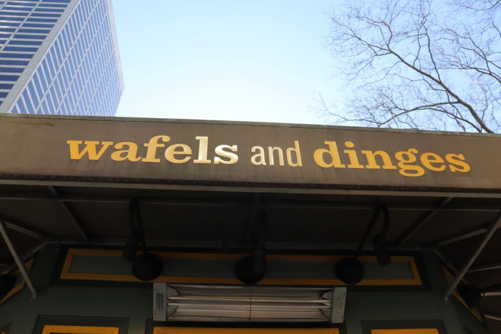 The sign for the wafels and dinges kiosk in NYC. There is light blue sky above and the top of a tree with a skyscraper in the upper left corner of the photo.