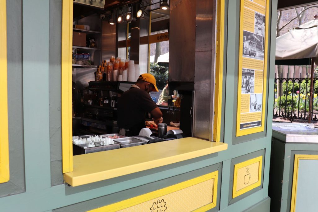 Looking in through the pick up window at the Wafels and Dinges kiosk in a New York City park. Inside there is a worker wearing a bright yellow ballcap,, preparing a waffle and there are stacks of containers and bins of utensils. The kiosk outer walls are muted green and medium yellow.
