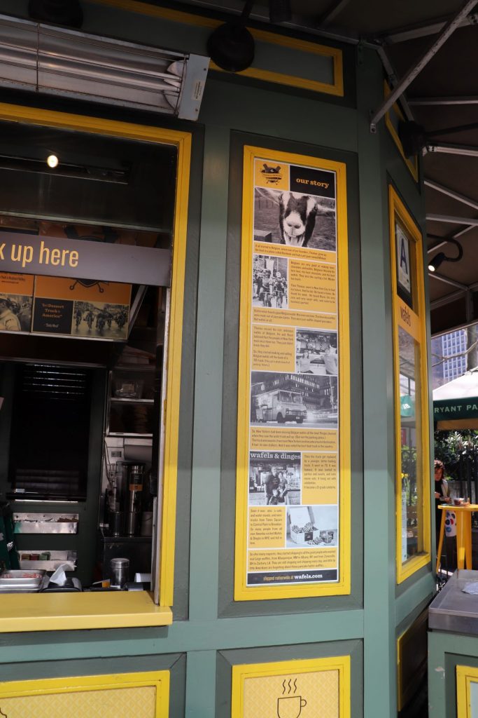 Side panel of the Wafels and Dinges kiosk at a New York City park. The walls are muted green and have yellow informational panels. To the left, is an open pick-up window.