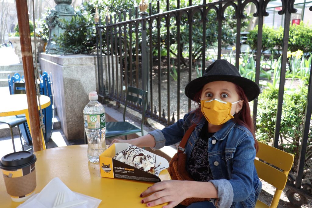 Anna, a teen girl sitting at patio seating at Wafels and Dinges in the park in New York City. She is wearing a denim jacket with a brown belt bag over her shoulder. She has on a black wide brim felt hat. She has an ice cream topped waffle in front of her and she is looking wide-eyed at the camera, as if astonished at how big of a size this treat is. There is also a coffee to-go cup and a water bottle on the table. She is wearing a bright yellow lower face mask.
