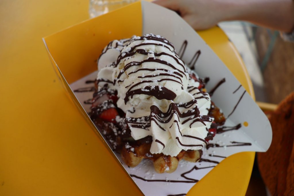 A waffle from Wafels and Dinges topped with strawberries, vanilla ice cream, whipped cream, choolate sauce drizzle and a sprinkle of powdered sugar. The to-go box is on top of a bright yellow patio table.