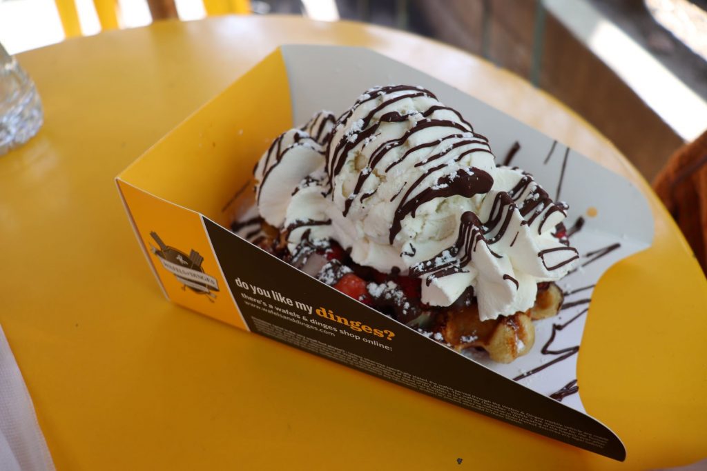 A waffle in an individual serving box topped with vanilla ice cream, whipped cream, powdered sugar sprinkles and chocolate sauce drizzle. The box is on a bright yellow patio table.