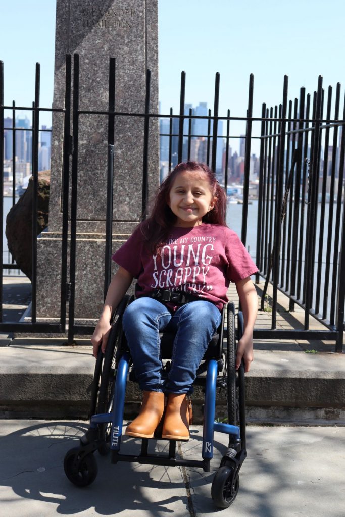 A view of the base of the Hamilton statue at the Weehawkin Dueling Grounds Anna, a teen girl wearing jeans and a t-shirt with the slogan "Young and Scrappy" is sitting in her wheelchair directly in front of the memorial. You can see a bit of the boulder behind the statue and there is a black wrought iron fence surrounding the site. Anna is smiling at the camera.