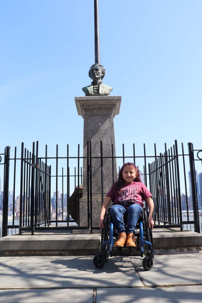 A view of the statue of Hamilton at the Weehawkin Dueling Grounds with the United States flag flying from a pole just behind. Anna, a teen girl wearing jeans and a t-shirt is sitting in her wheelchair directly in front of the memorial. You can see the curb cut ramp directly in front of her leading to the street.