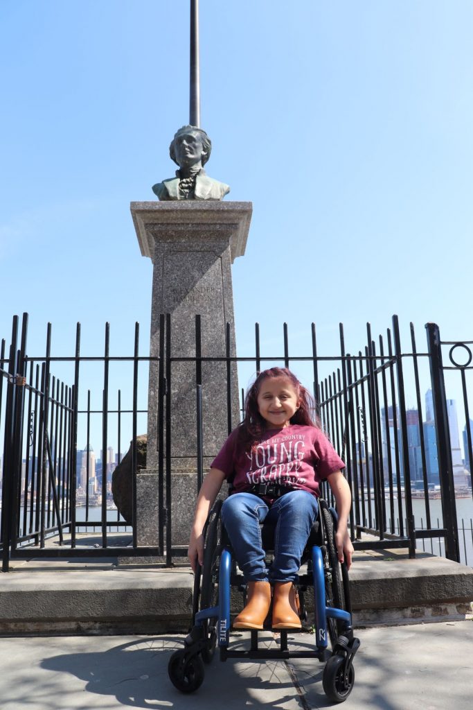 A view of the statue of Hamilton at the Weehawkin Dueling Grounds with a flag pole just behind. Anna, a teen girl wearing jeans and a t-shirt is sitting in her wheelchair directly in front of the memorial. You can see the curb cut ramp directly in front of her leading to the street.