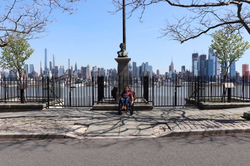 A view of the statue of Hamilton at the Weehawkin Dueling Grounds with a flag pole just behind. Anna, a teen girl wearing jeans and a t-shirt is sitting in her wheelchair directly in front of the memorial. You can see the curb cut ramp directly in front of her leading to the street. The river and a view of NYC is in the background.