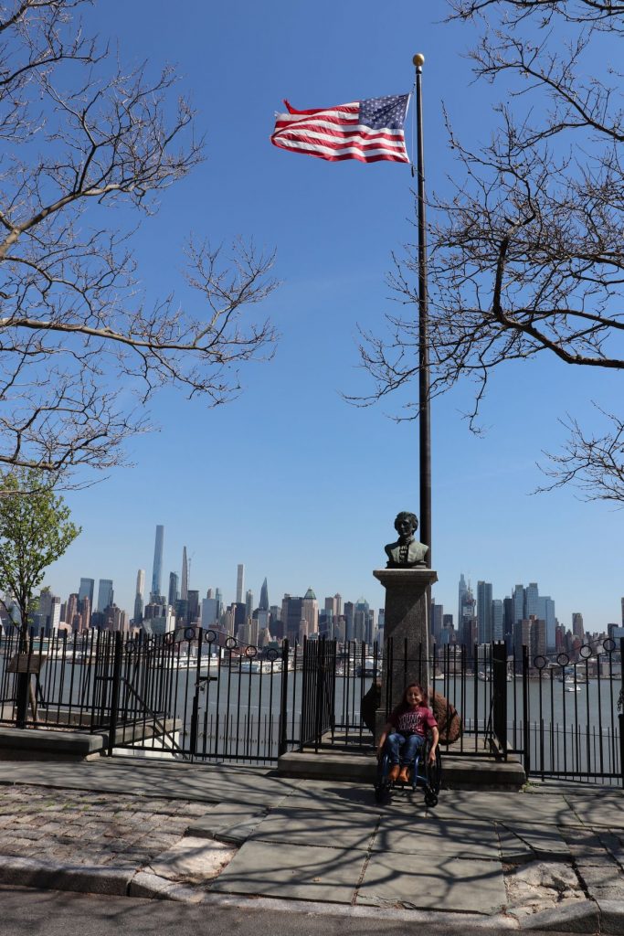 A view of the statue of Hamilton at the Weehawkin Dueling Grounds with the United States flag flying from a pole just behind. Anna, a teen girl wearing jeans and a t-shirt is sitting in her wheelchair directly in front of the memorial. You can see the curb cut ramp directly in front of her leading to the street.