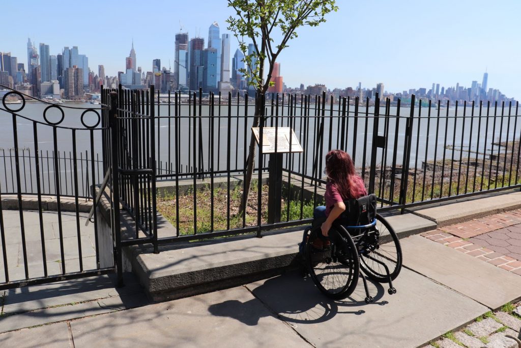 Close up photo showing Anna,, a teen girl using a wheelchair, reading a historic information sign at the Weehawking Dueling Grounds. One arm is by the side of the chair and she has long hair and is wearing a t-shirt. The sign is surrounded by an iron fence and the river and city of NYC are in the background.