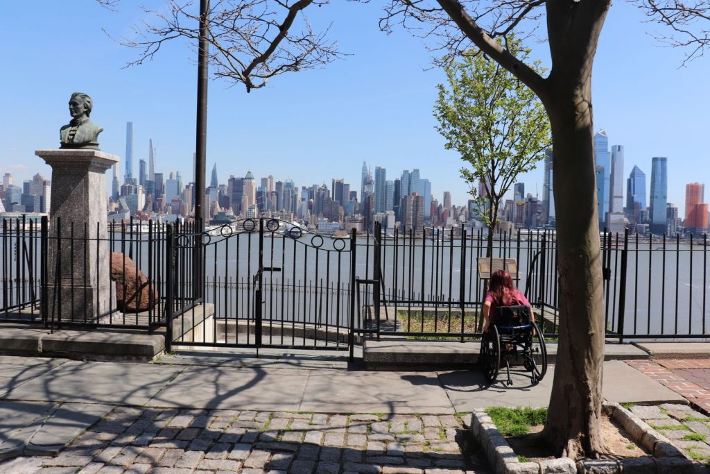 Anna, a teen girl and huge fan of the Hamilton musical, is reading an informational sign posted at the Weehawkin Dueling Grounds. Her back is to the camera and you see her long hair, her burgundy heather Hamilton t-shirt and she is sitting in her wheelchair. The river and skyline of NYC are in the background.
