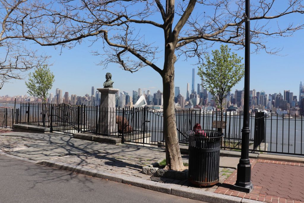 A street view showing ramp access to the Alexander Hamilton statue at Weehawken Dueling Grounds. There is the sidewalk in front with the wrought iron fence surrounding the memorial and a black metal trash can and lamp post in the lower right corner. The river and skyline of NYC are visible in the background.